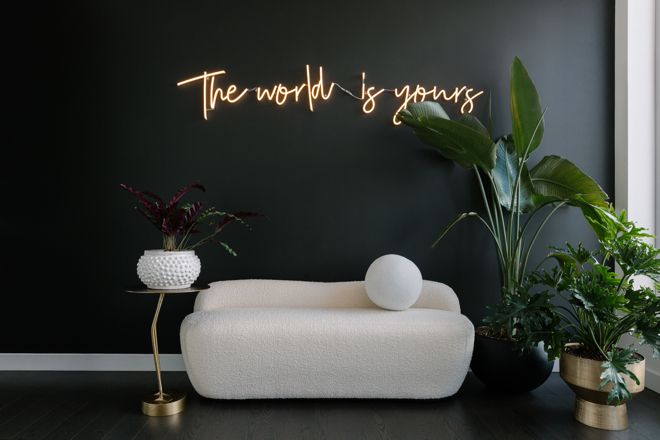 White loveseat and plants against black wall with neon sign